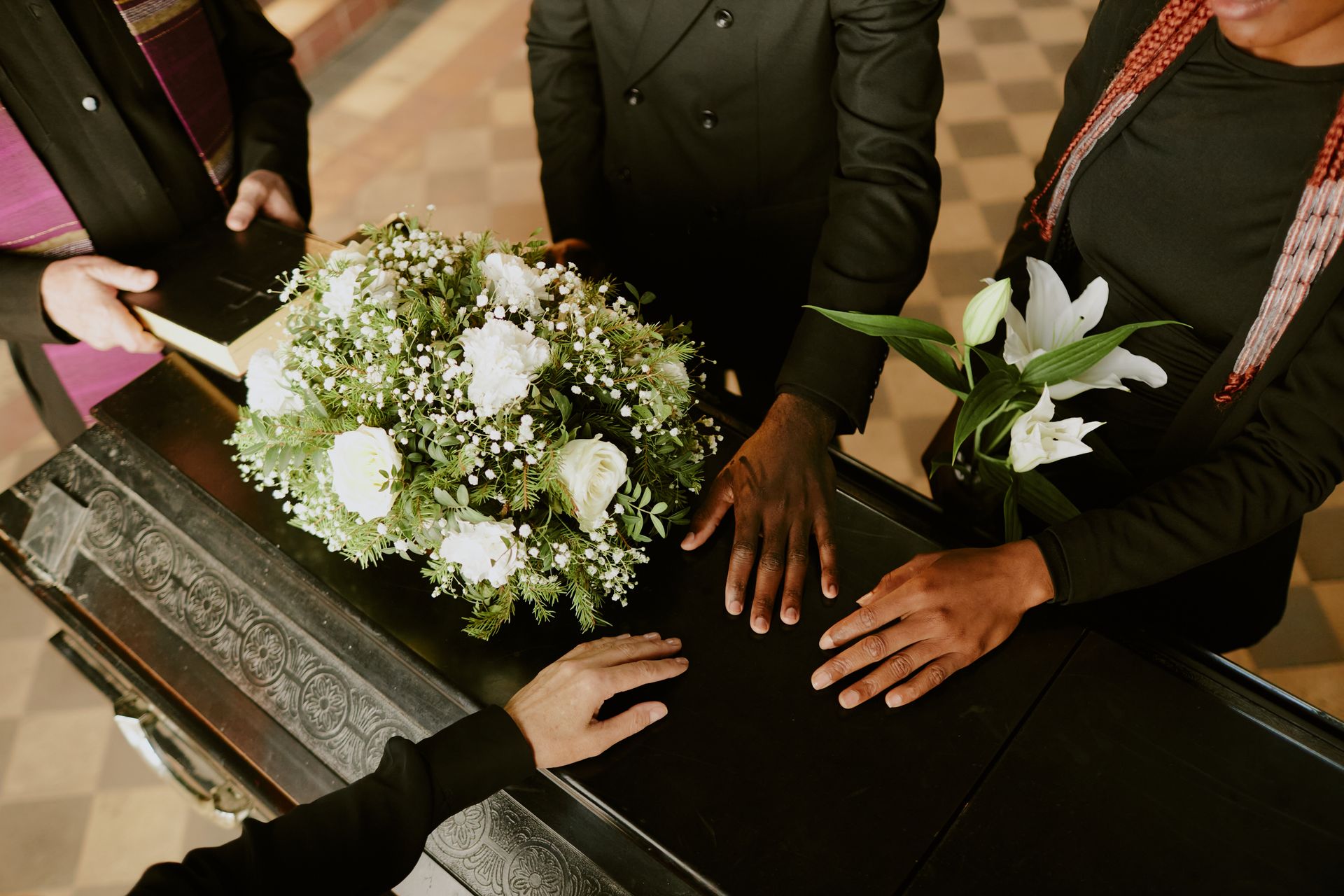 People in dark clothing gather around a casket adorned with white flowers, placing their hands on it in a moment of grief.