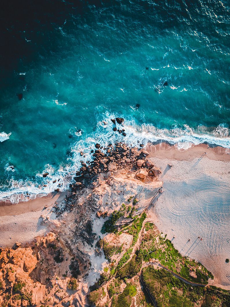 Aerial view of rocky, grassy cliffs meeting a sandy beach and vibrant turquoise ocean with white crashing waves.