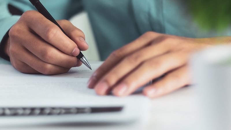 Hands resting on a desk, with one hand holding a pen while writing on a document.