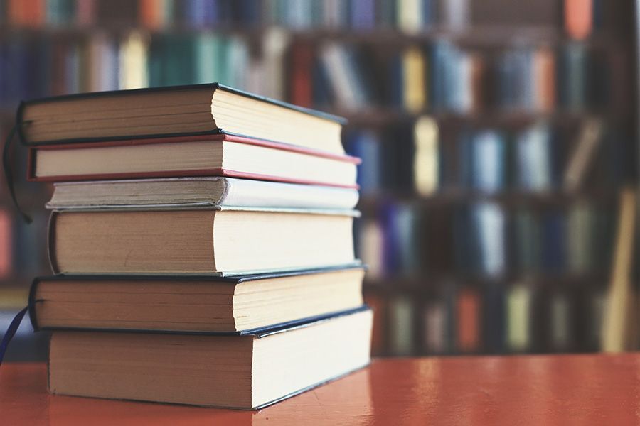 A stack of five hardcover books rests on a table, with a blurred bookshelf in the background.
