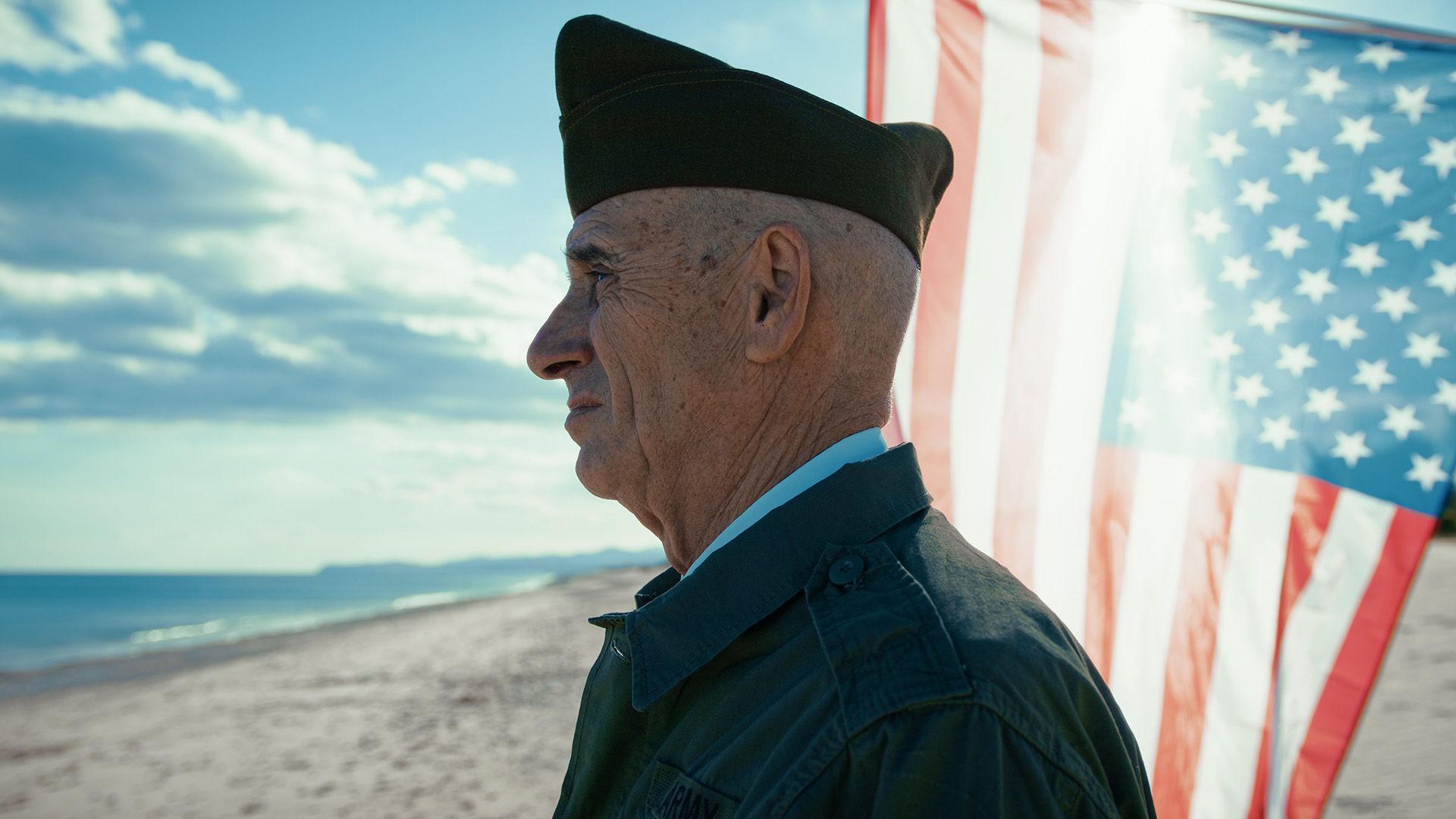 A veteran in uniform stands on a beach in profile, with a large American flag waving beside them against a sunny sky.