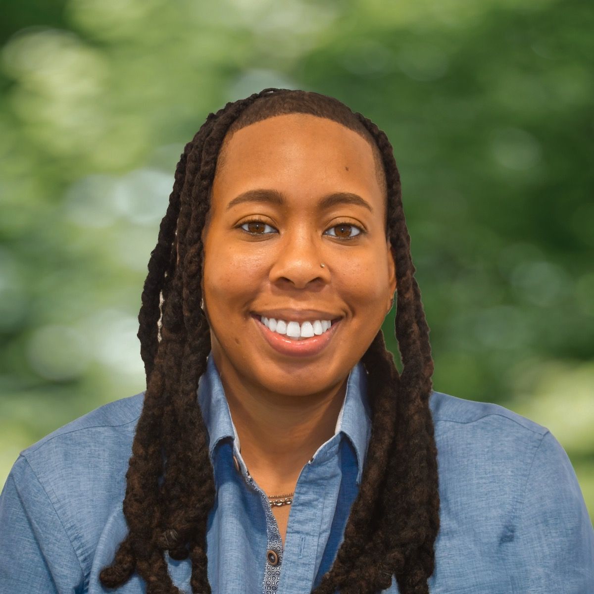 Man with dark hair and beard, wearing striped shirt, smiling, and gazing at the camera, in a bright setting.