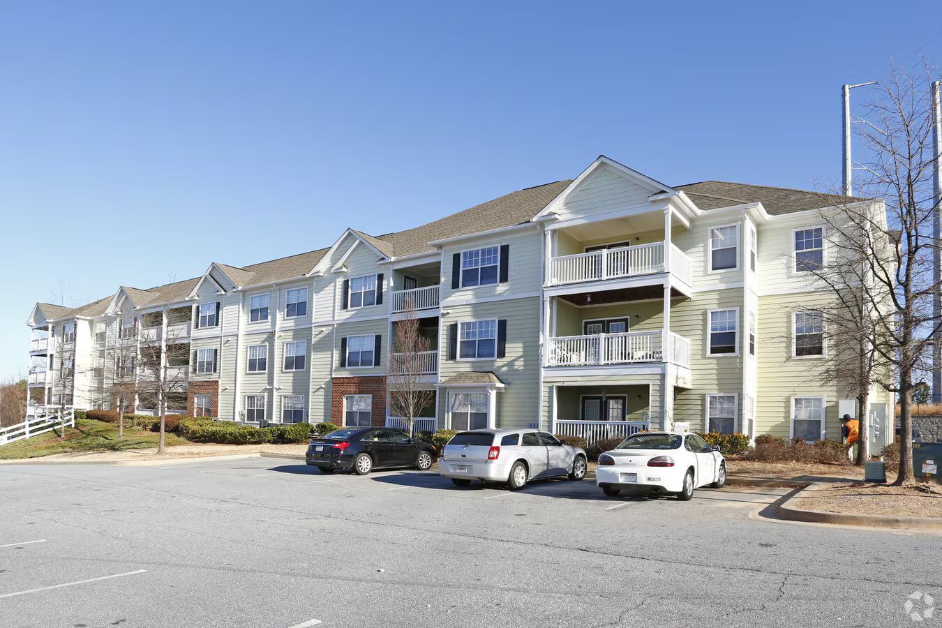 A three-story apartment complex with beige siding and a parking lot in front under a clear blue sky.