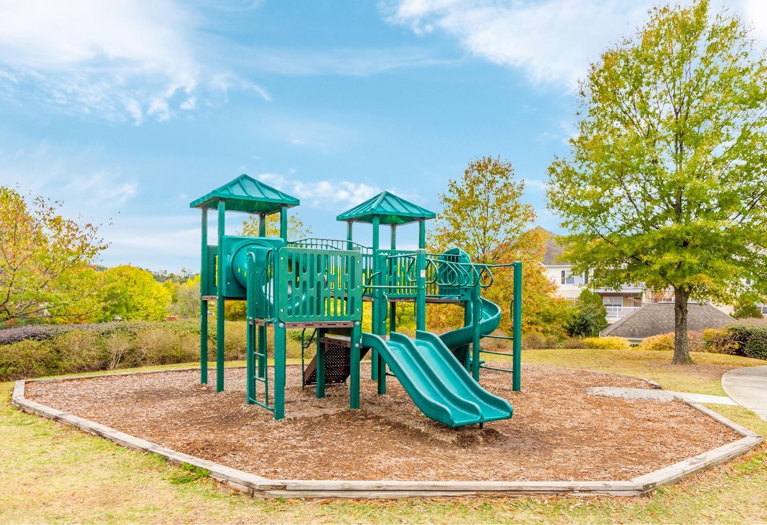 A green playground structure with slides and a tube tunnel, surrounded by mulch and trees under a partly cloudy sky.