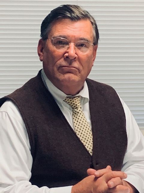 Man in glasses, brown vest, and yellow-patterned tie, seated with clasped hands; neutral expression.