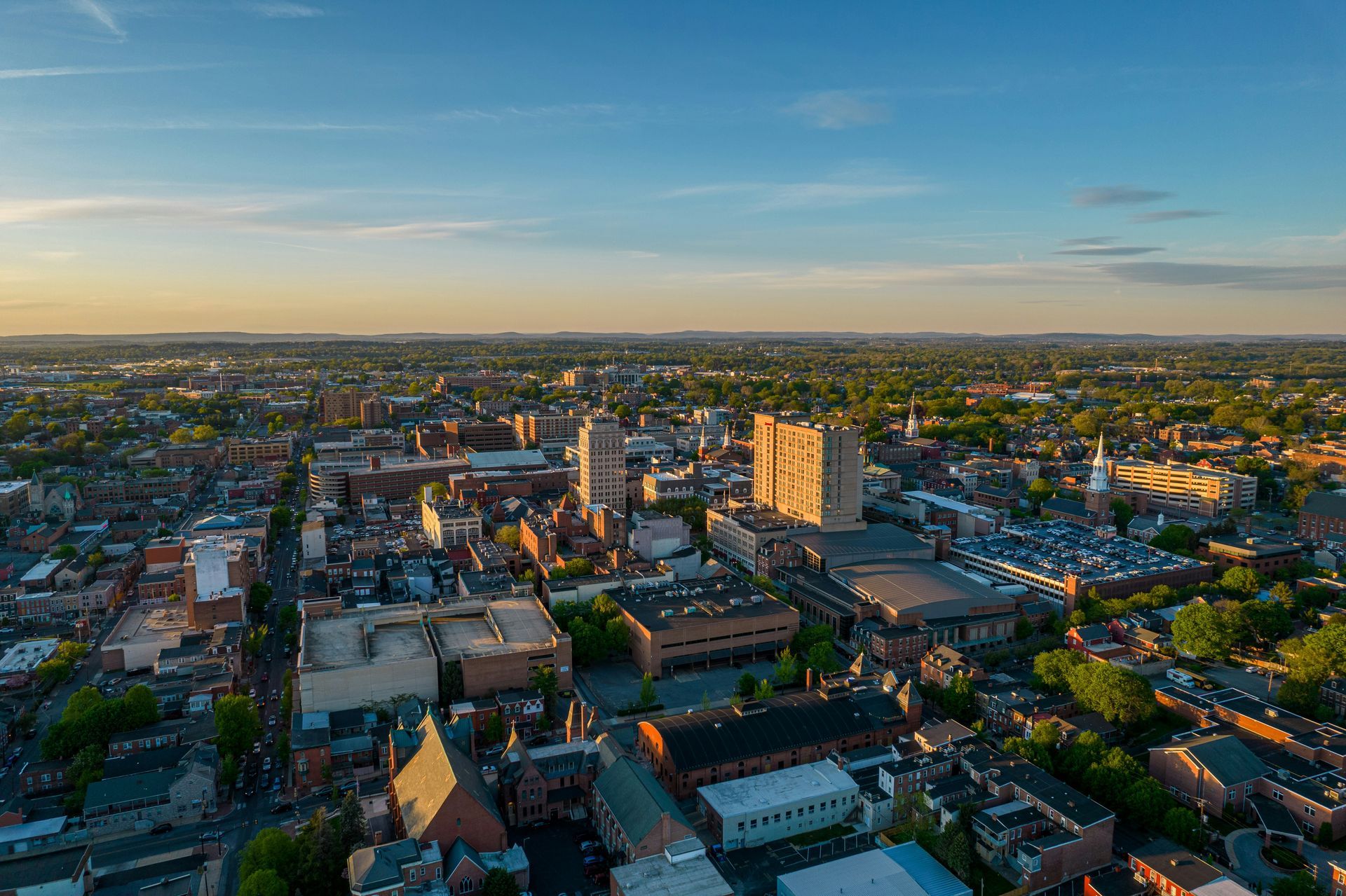 Aerial view of a city with buildings and trees under a clear, blue sky at sunset.