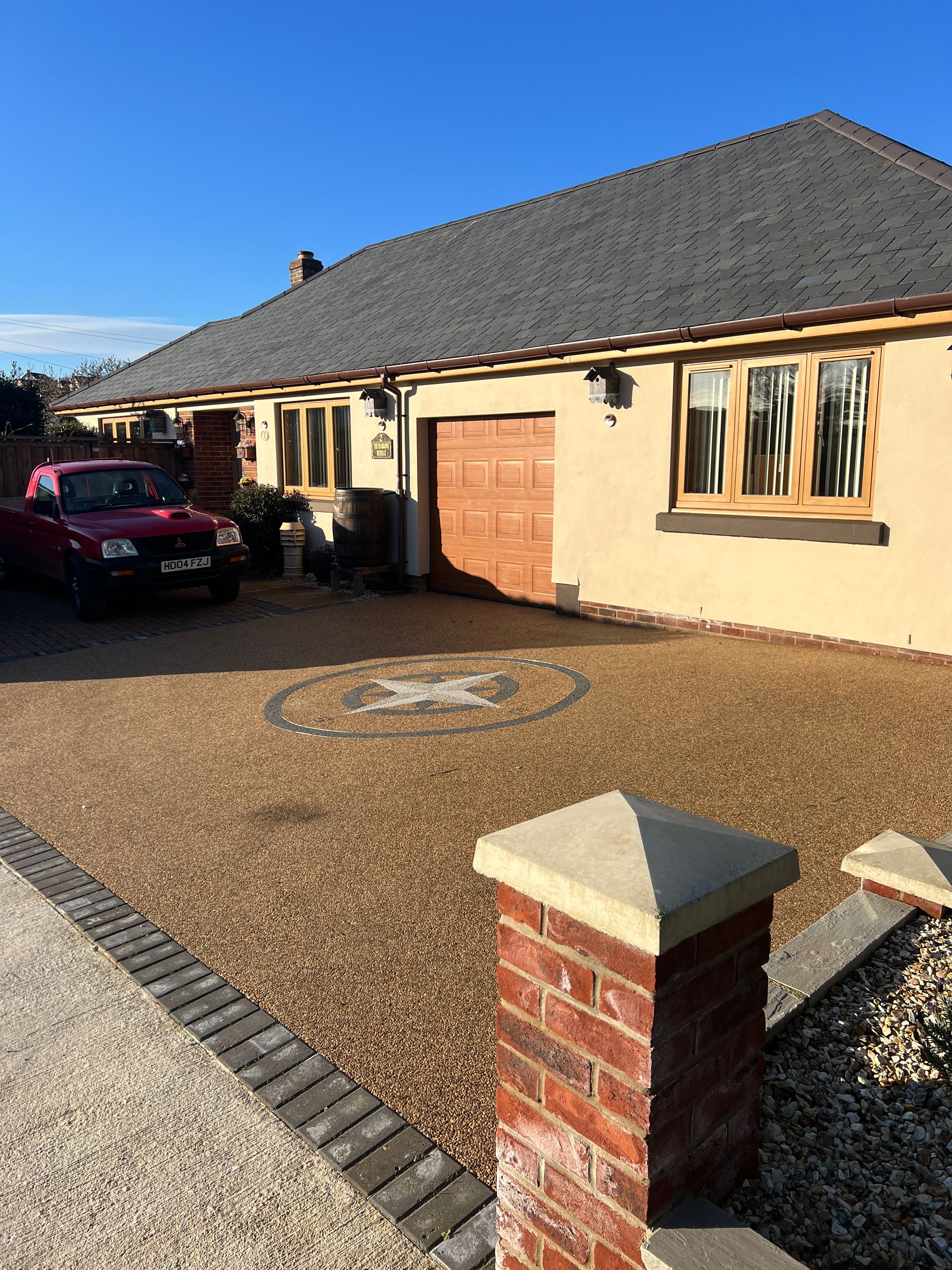 Brown resin driveway with a compass pattern on it, in front of a cream house with a brown garage.