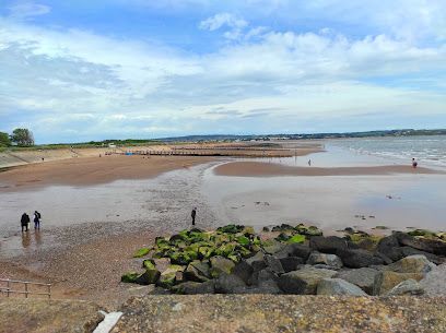 Beach scene with sand, water, and cloudy sky; people walking; rocks in foreground.