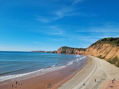Beach scene: blue sky, ocean, sand, red cliffs, people walking.