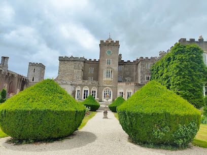 A stone castle with a central clock tower, framed by trimmed green bushes along a gravel path under a cloudy sky.