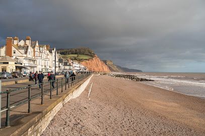 Beachfront promenade with buildings, people, and a sandy beach under a cloudy sky.