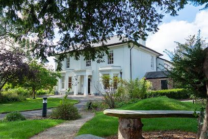 White house with columns, garden, stone bench, and trees under blue sky.