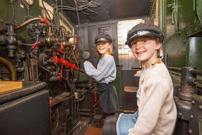 Two children in train engineer hats smile inside a train cab, surrounded by controls.