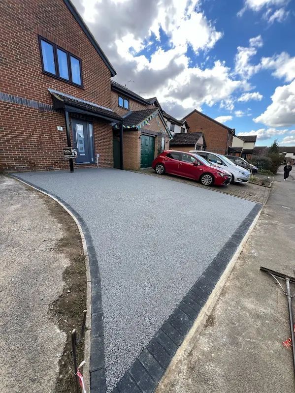 A newly paved gray driveway with black borders in front of a brick house with cars parked alongside it.