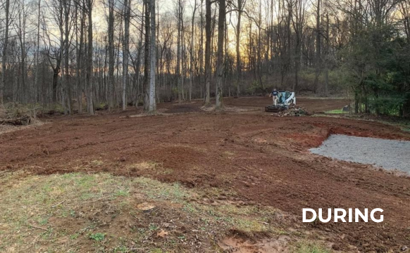 A dirt field with trees in the background and a bulldozer in the middle of it. DURING PICTURE FOR GRADING PROJECT. 