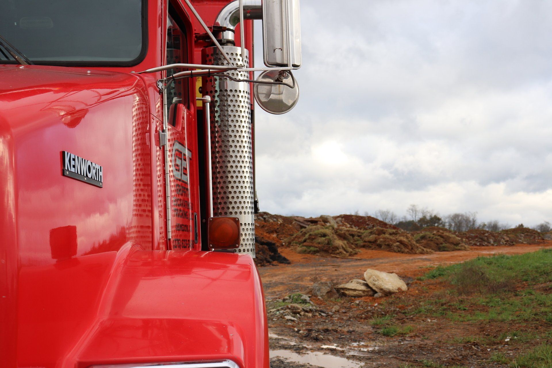 A red truck is parked in a field with a pile of wood in the background.