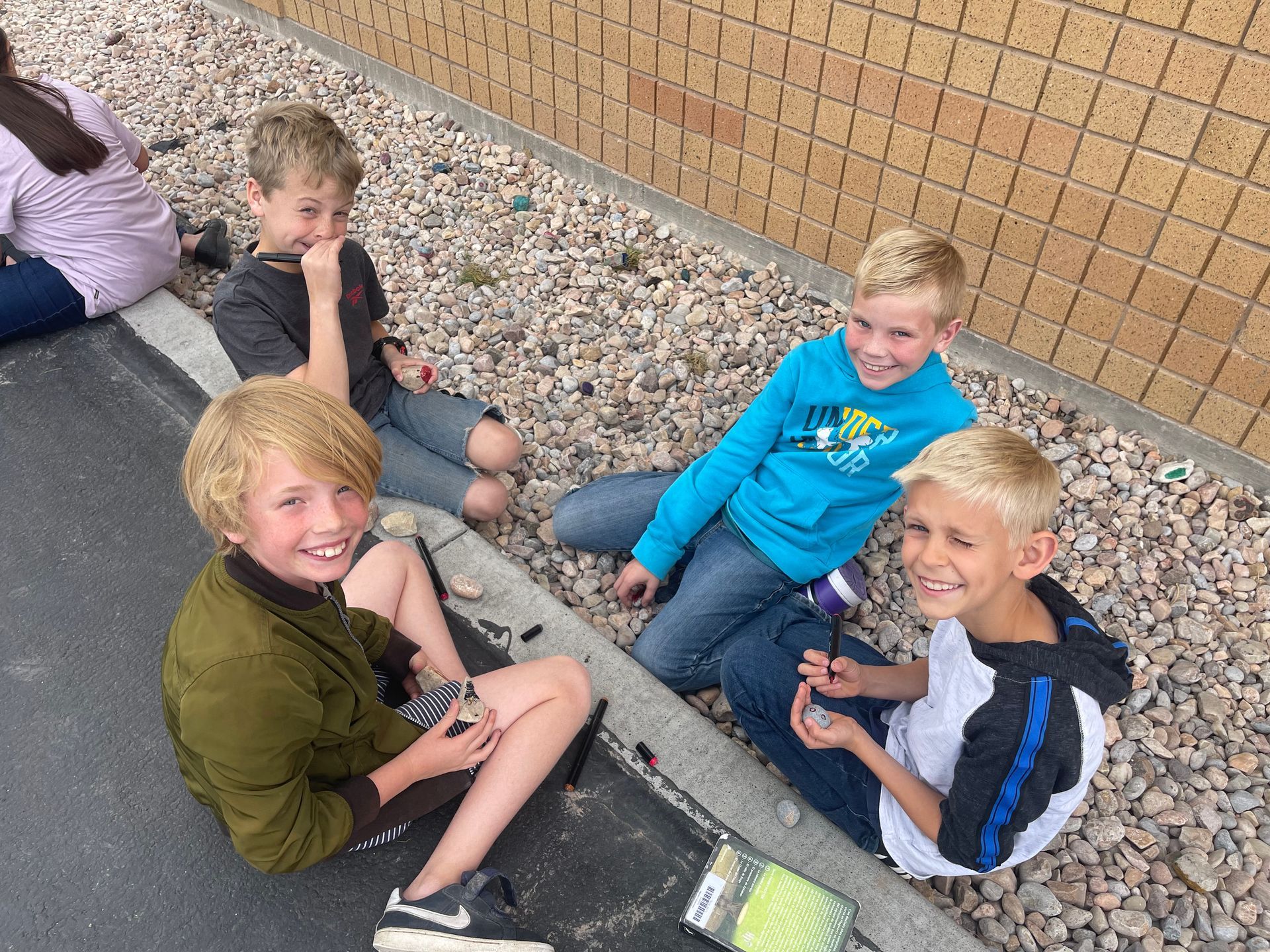 Fifth grade students smiling while they paint rocks on the ground outside the school.