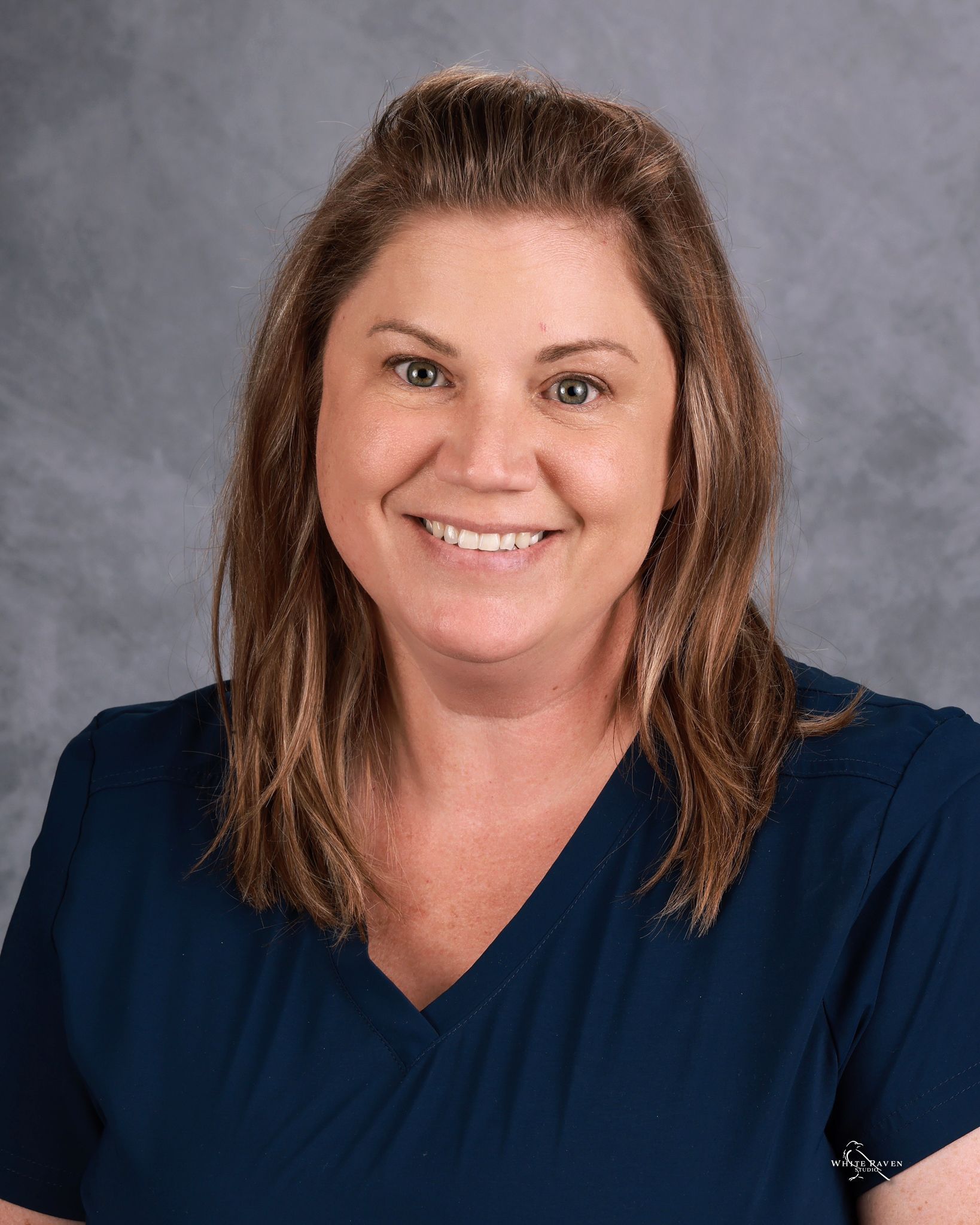 A person with shoulder-length brown hair, wearing a navy blue V-neck top, smiling against a mottled grey background.