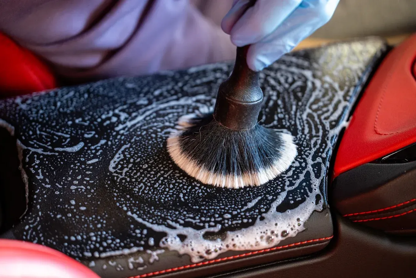 Gloved hand using a brush to scrub foamy cleaner on black car interior with red accents.