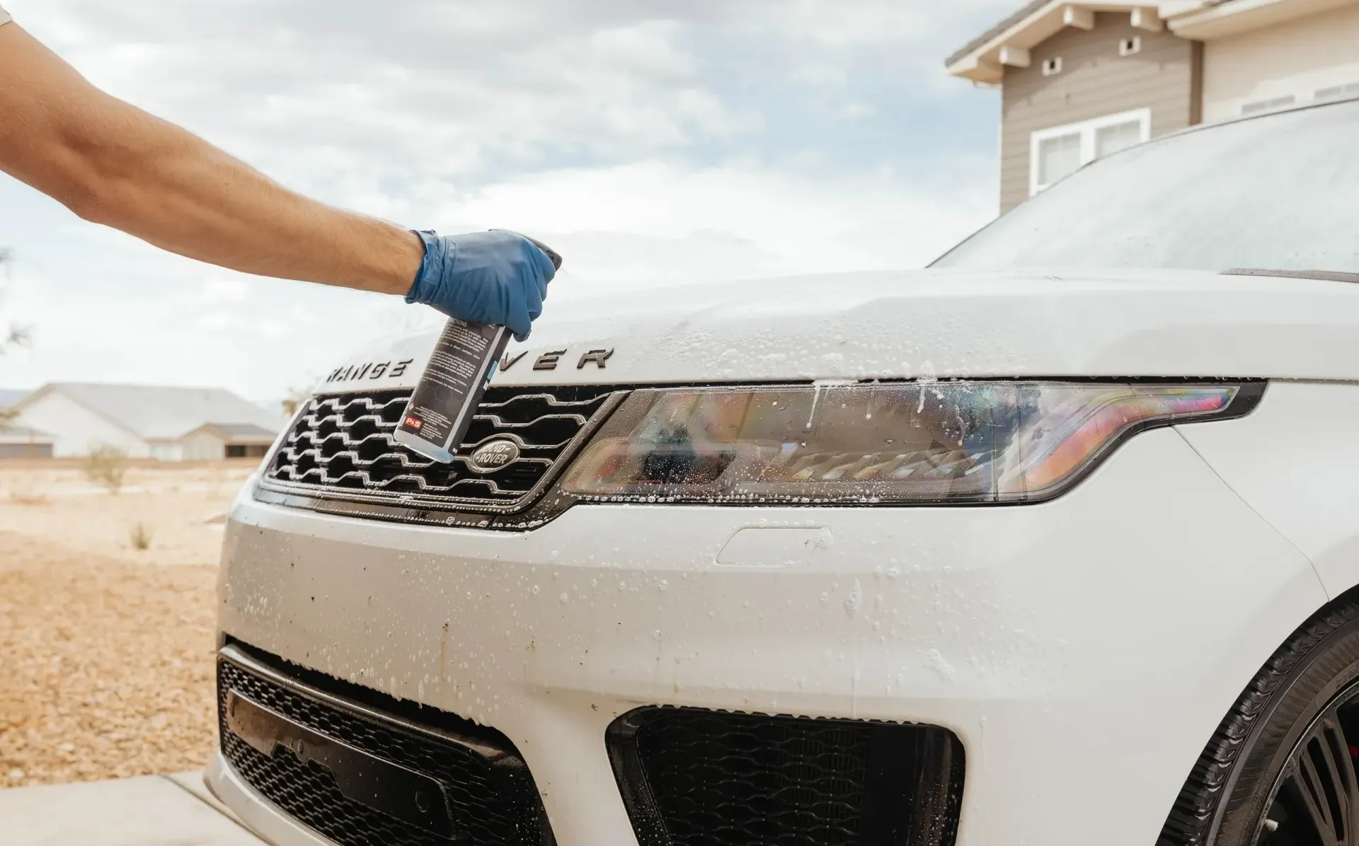 A person in blue gloves sprays cleaning product on the front of a white Range Rover SUV.