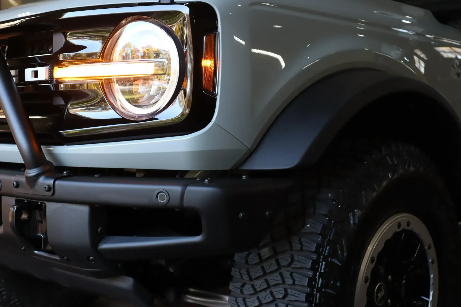 Close-up of a light gray Ford Bronco's front end, showing a round headlight, turn signal, and black bumper.