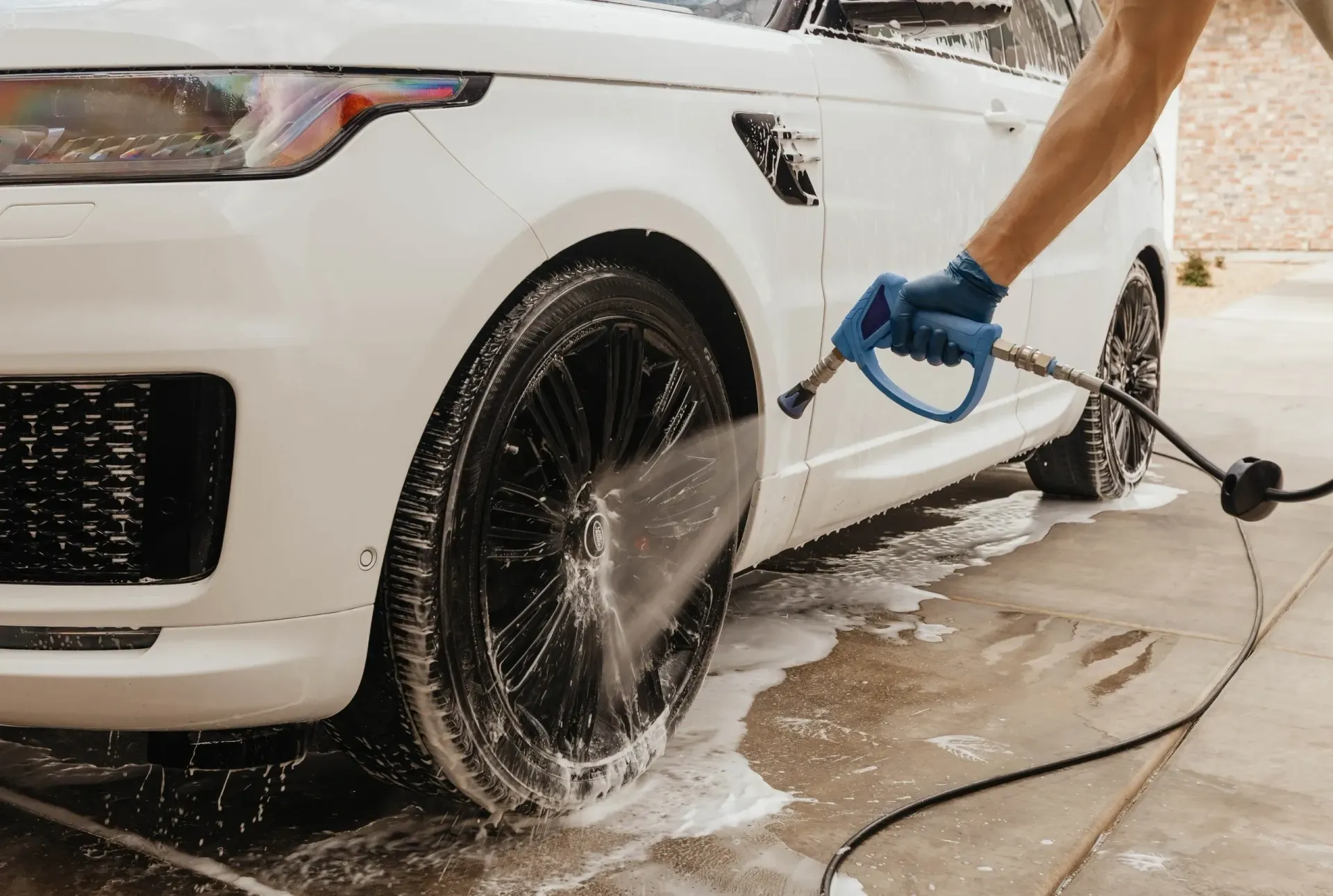 A person wearing a blue glove sprays a white car's black tire with soapy water.