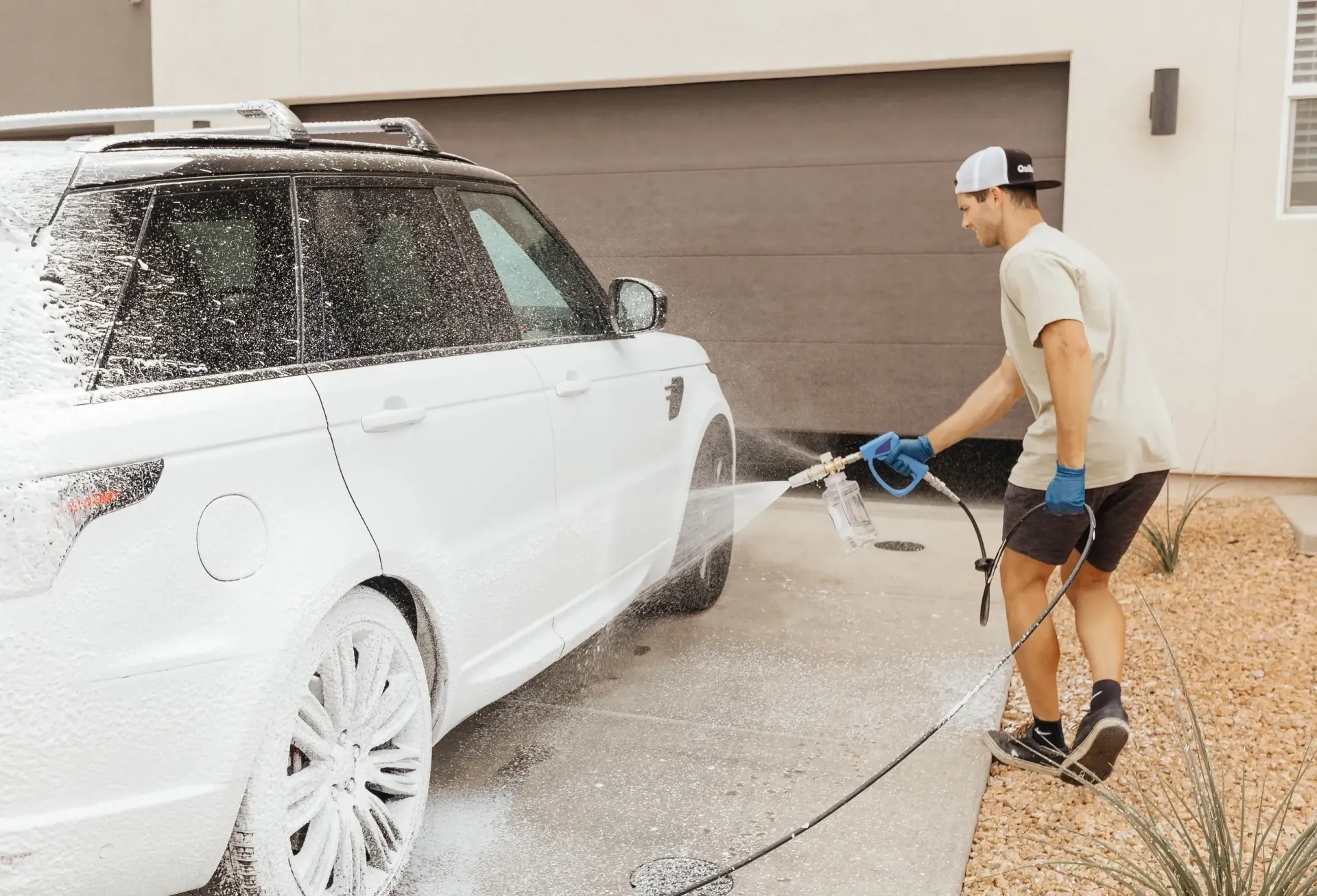 Man washing a white SUV with soapy foam in front of a house.
