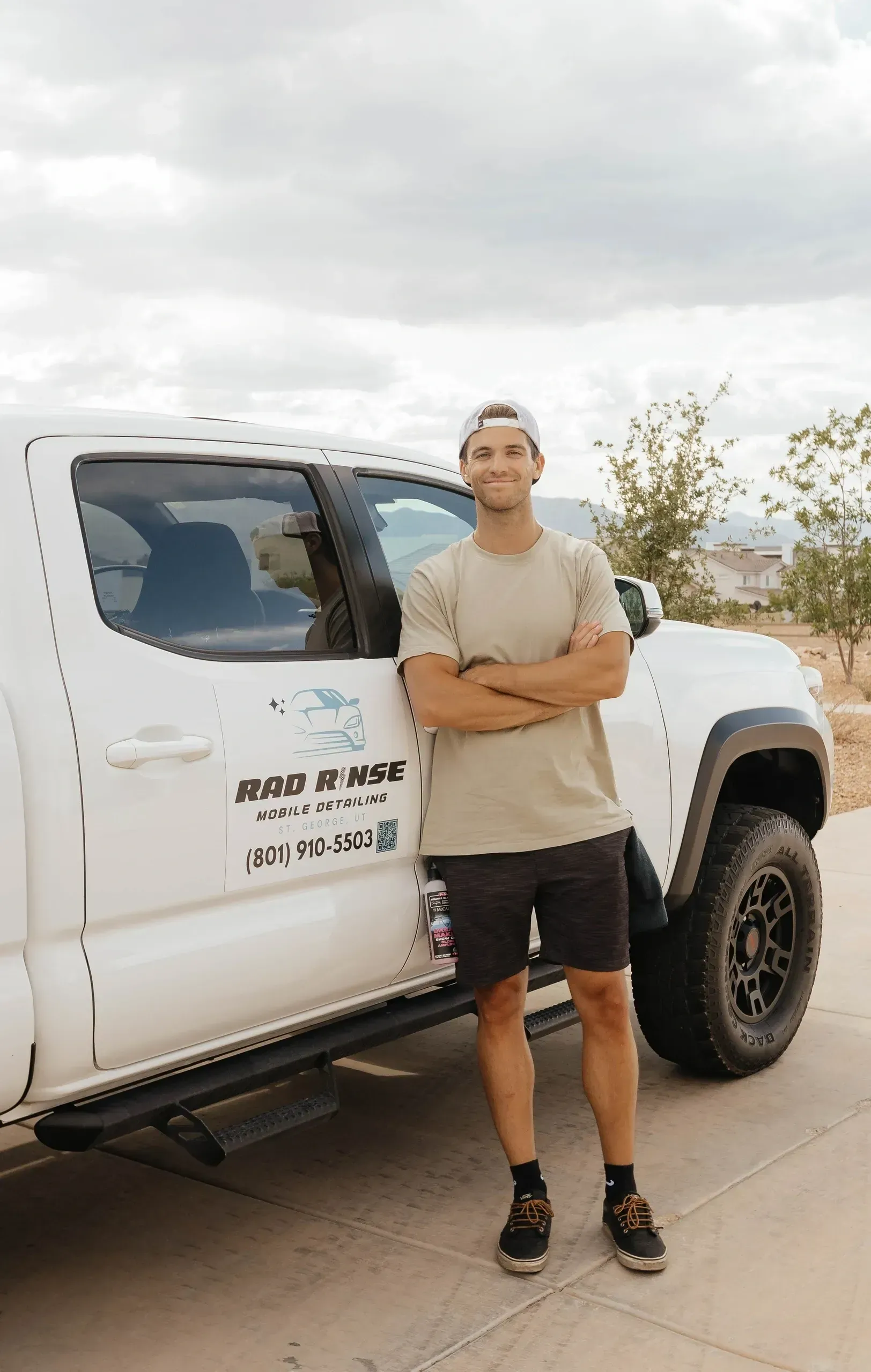 Man standing with arms crossed next to a white truck with business logo; outdoors.