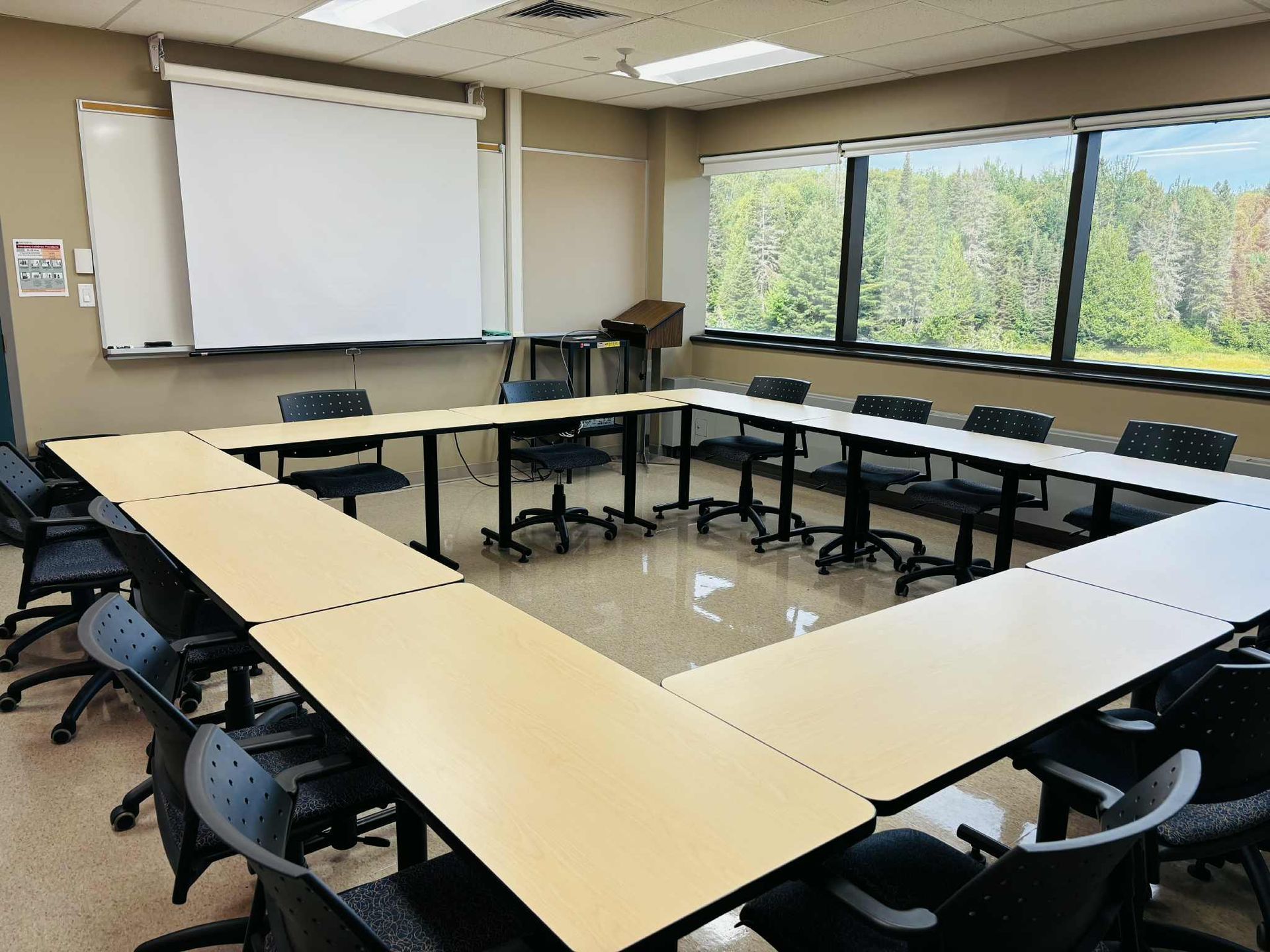 Classroom with tables arranged in a square, chairs around tables, windows, whiteboard, and projector screen.