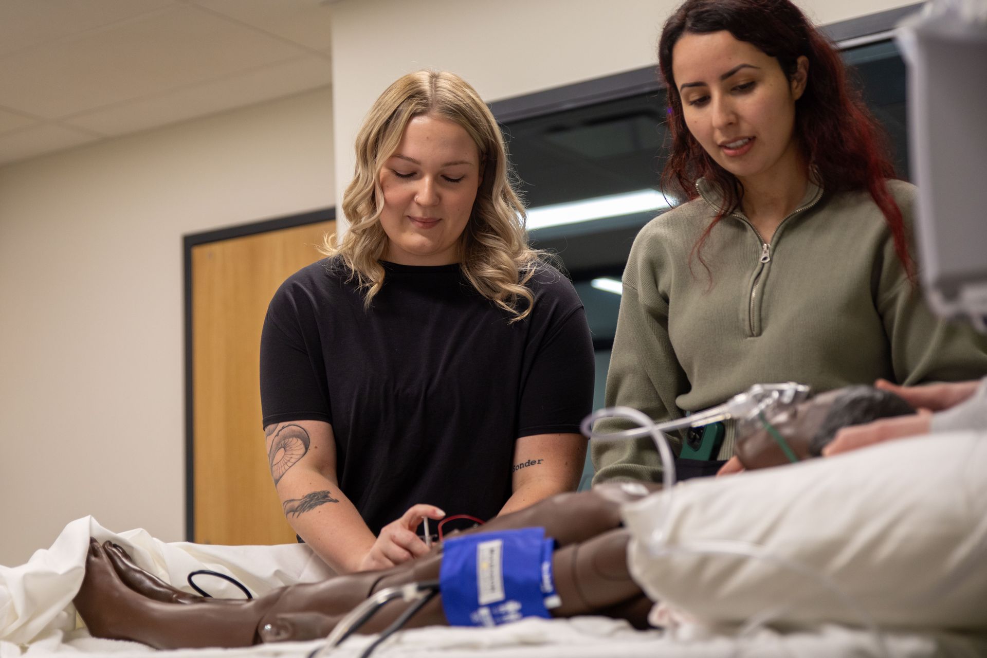 Two women are standing next to a mannequin on a bed.