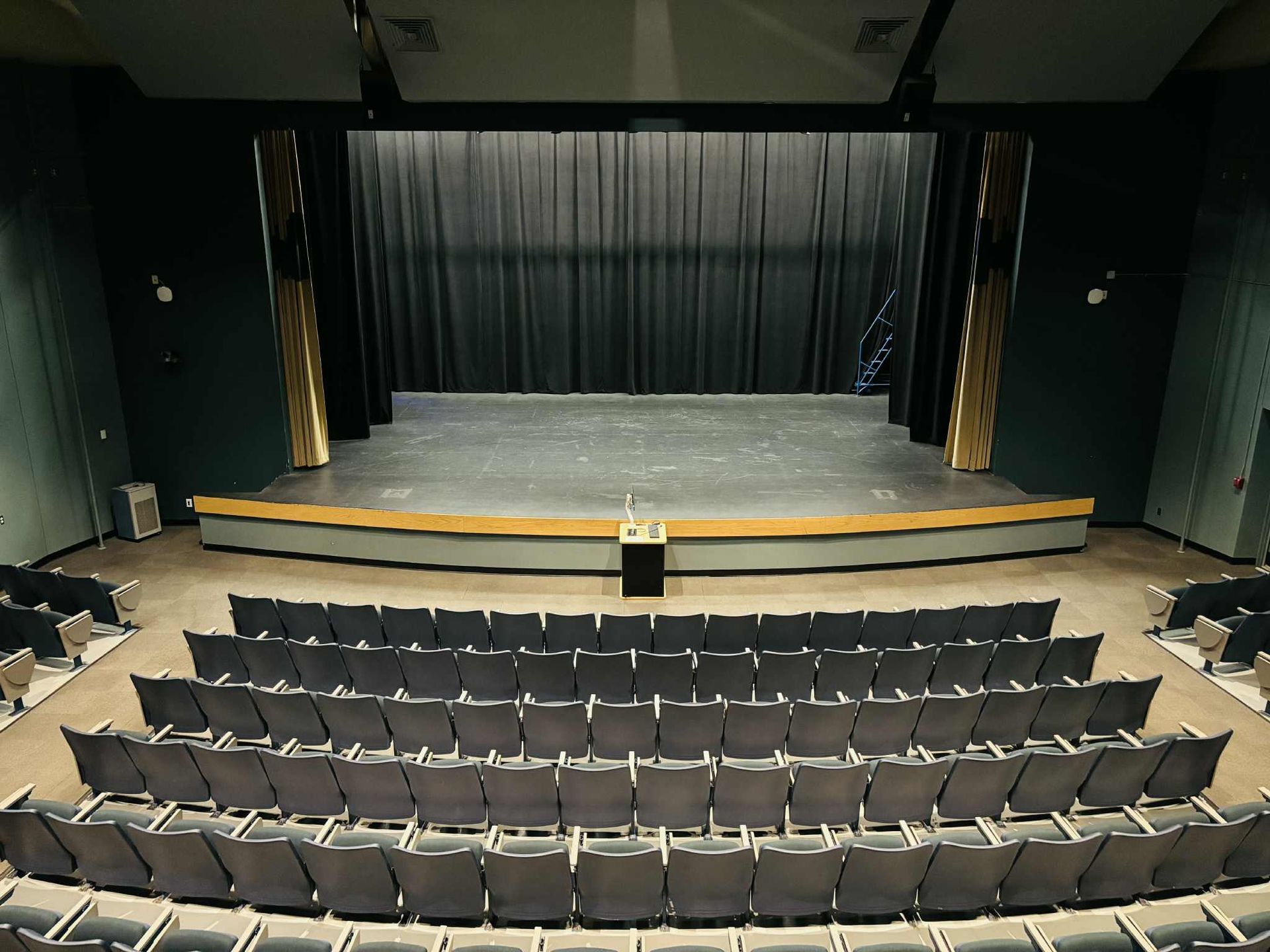 Rows of theater seats face a stage with black curtains, a podium, and gold-trimmed columns.