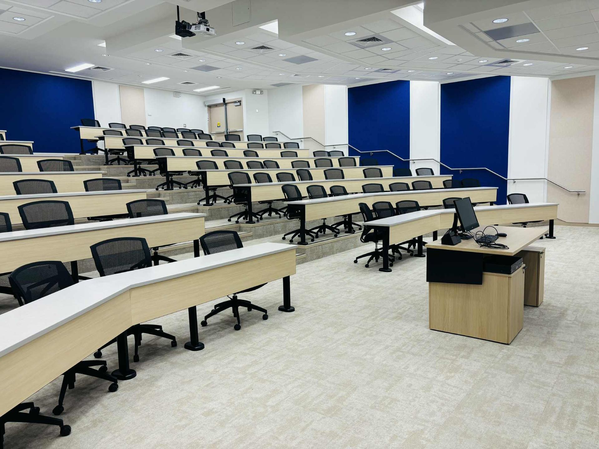 Empty tiered lecture hall with long desks, projector screen, and rows of chairs.