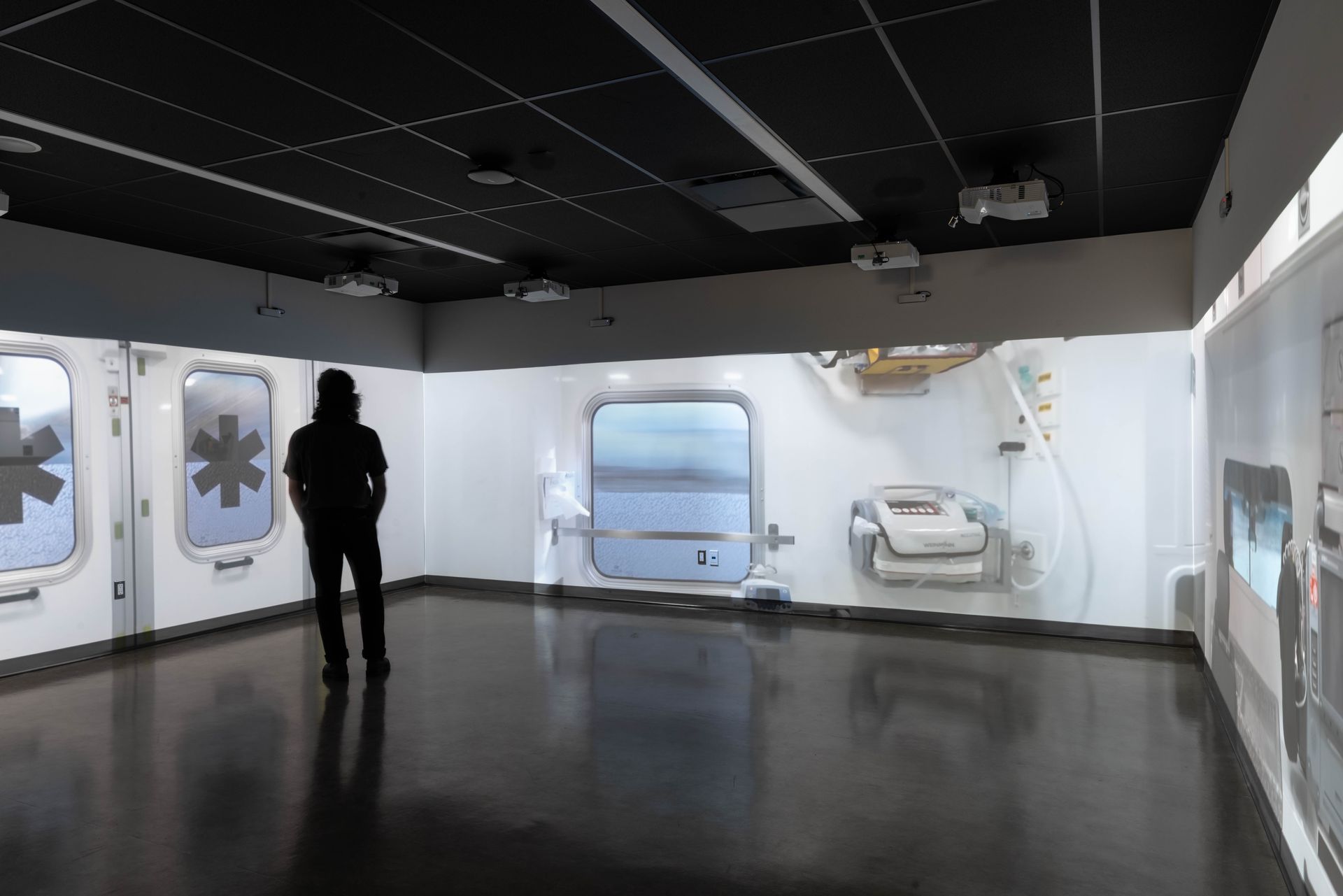 A person stands in a white room with medical symbols. The walls have windows and equipment.