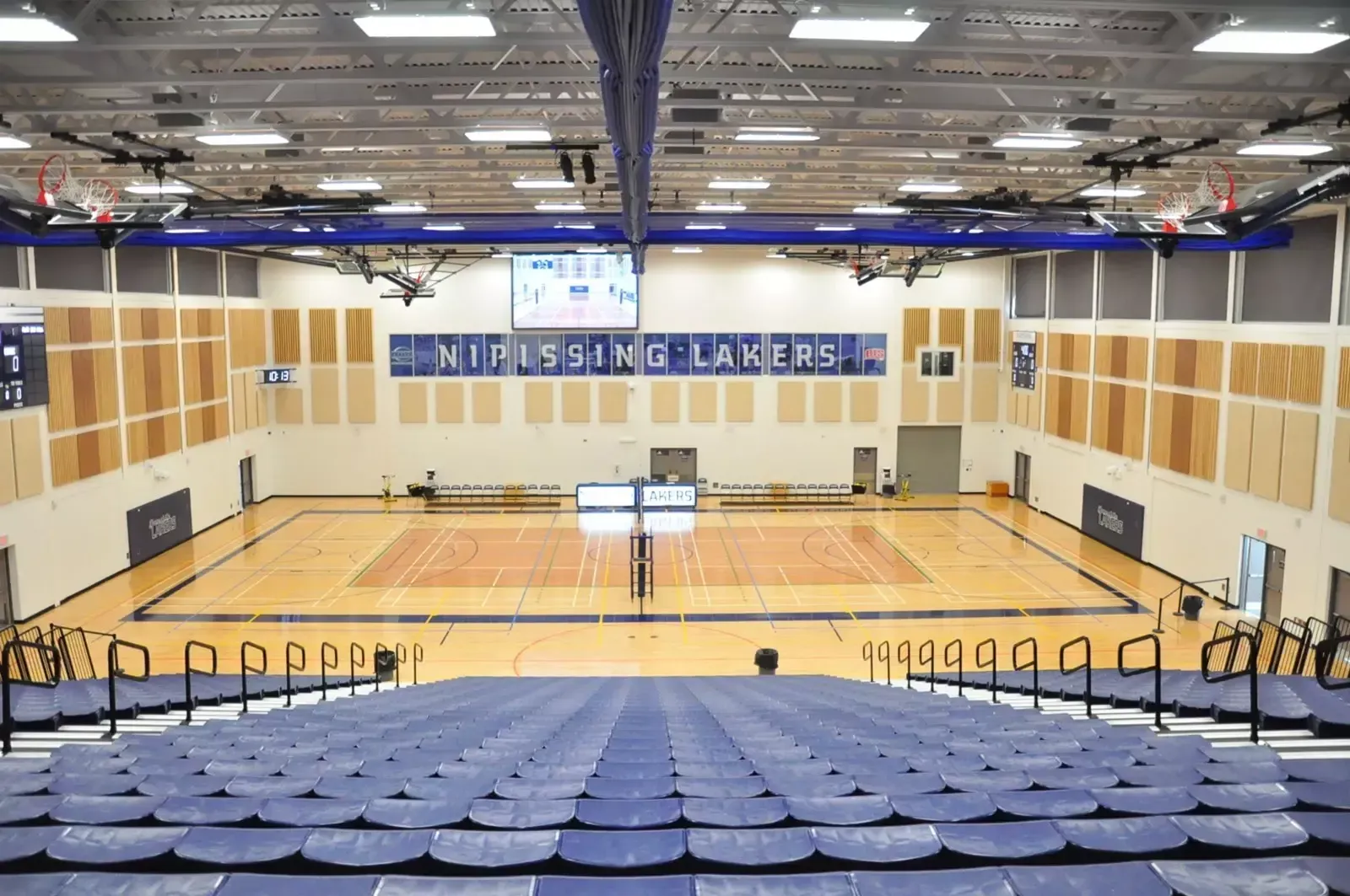 Nipissing University gym, large crowd watches from bleachers. Players in action on court, blue and white color scheme.
