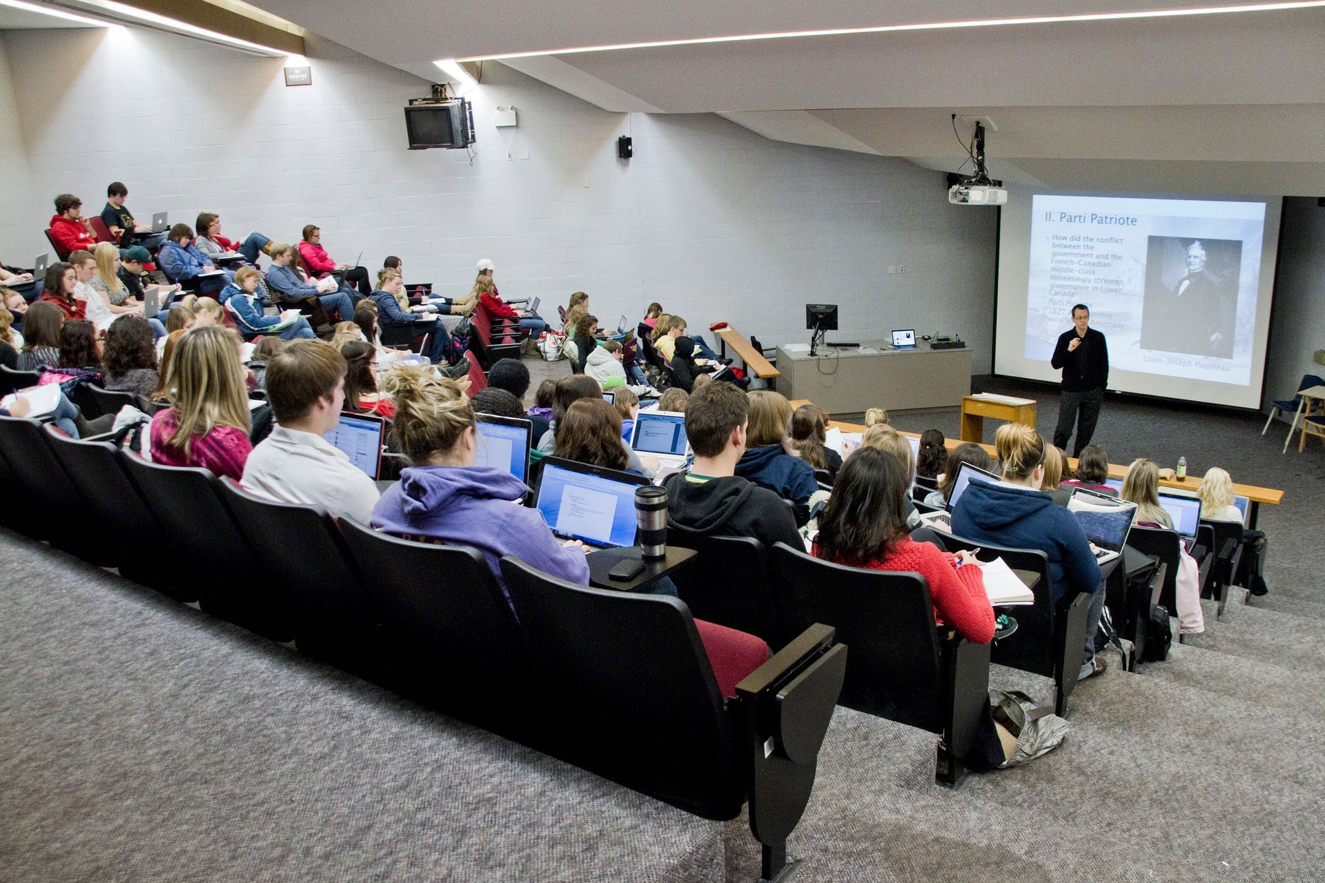 A professor lectures to students in a tiered lecture hall. Students are working on laptops.