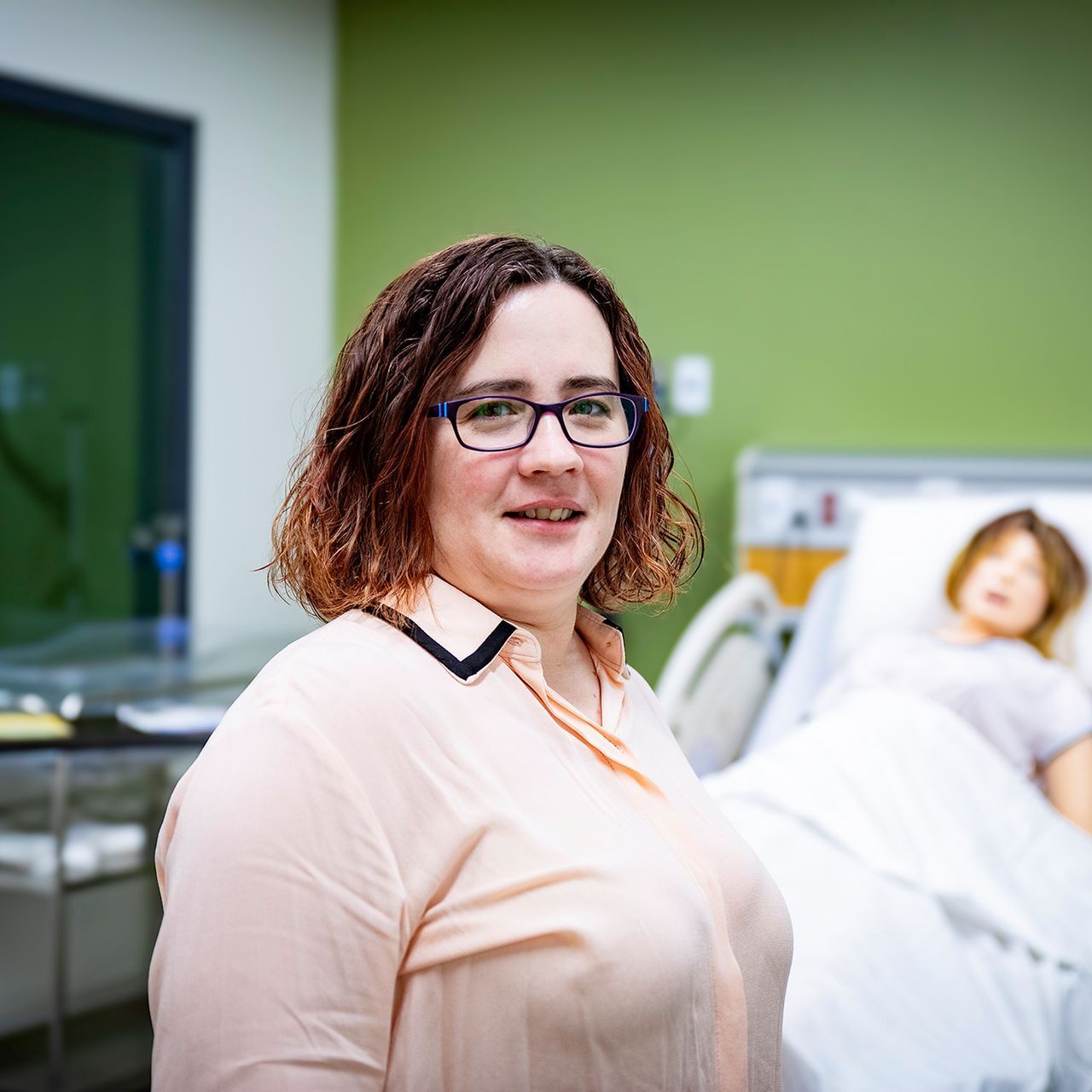 A woman wearing glasses is standing in front of a hospital bed.