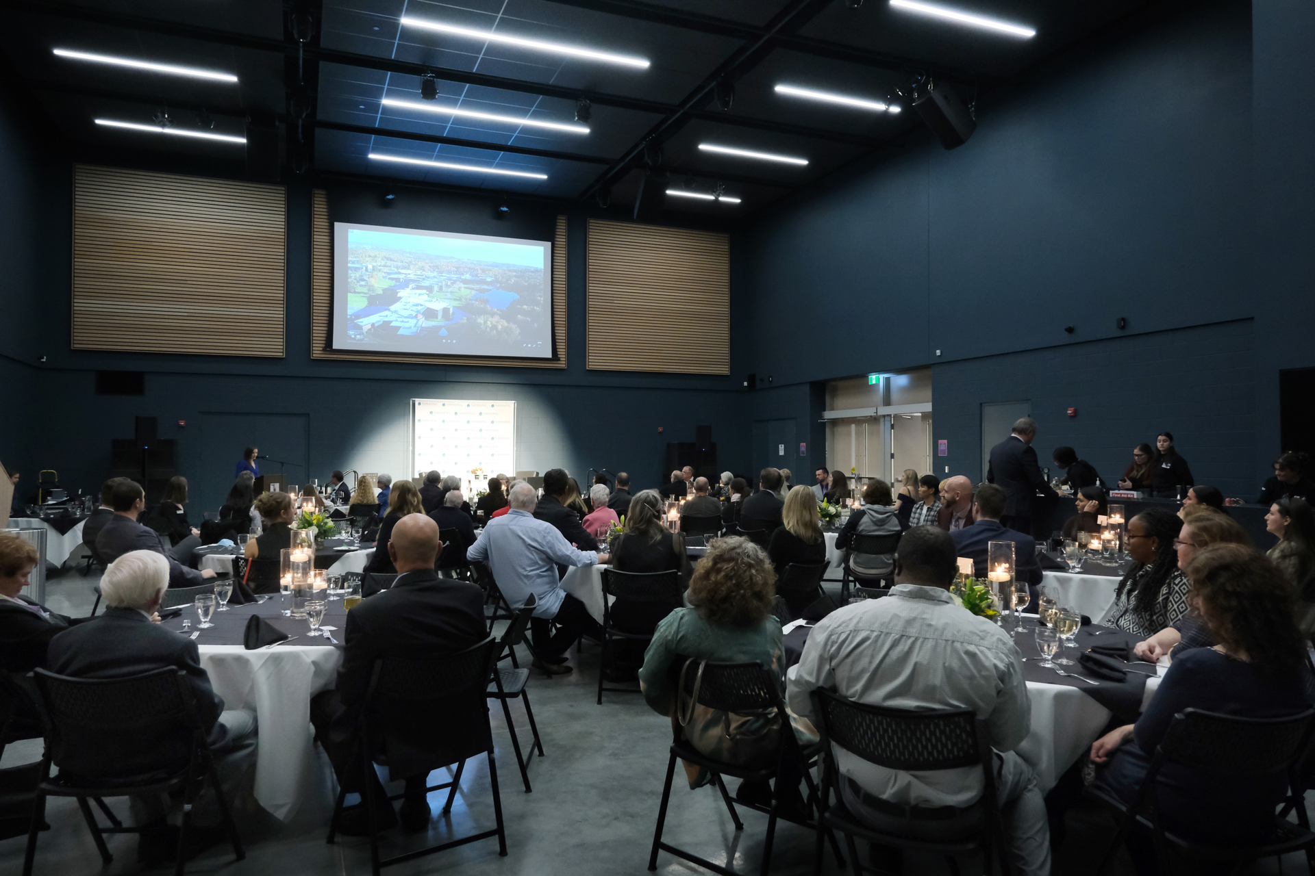 An event in a large hall: people seated at tables, a screen displaying an image, and architectural details.