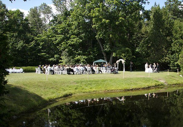 Wedding ceremony by a pond, guests seated, white arch, green grass and trees.