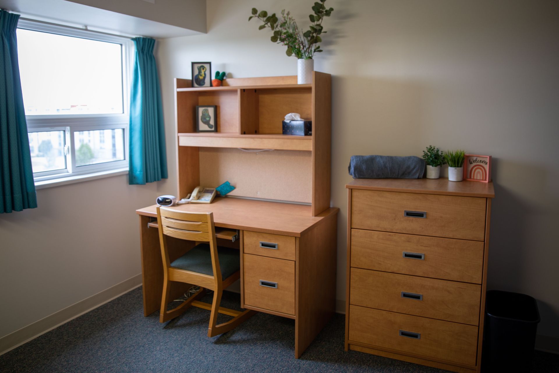 A bedroom with a desk , chair , dresser and window.