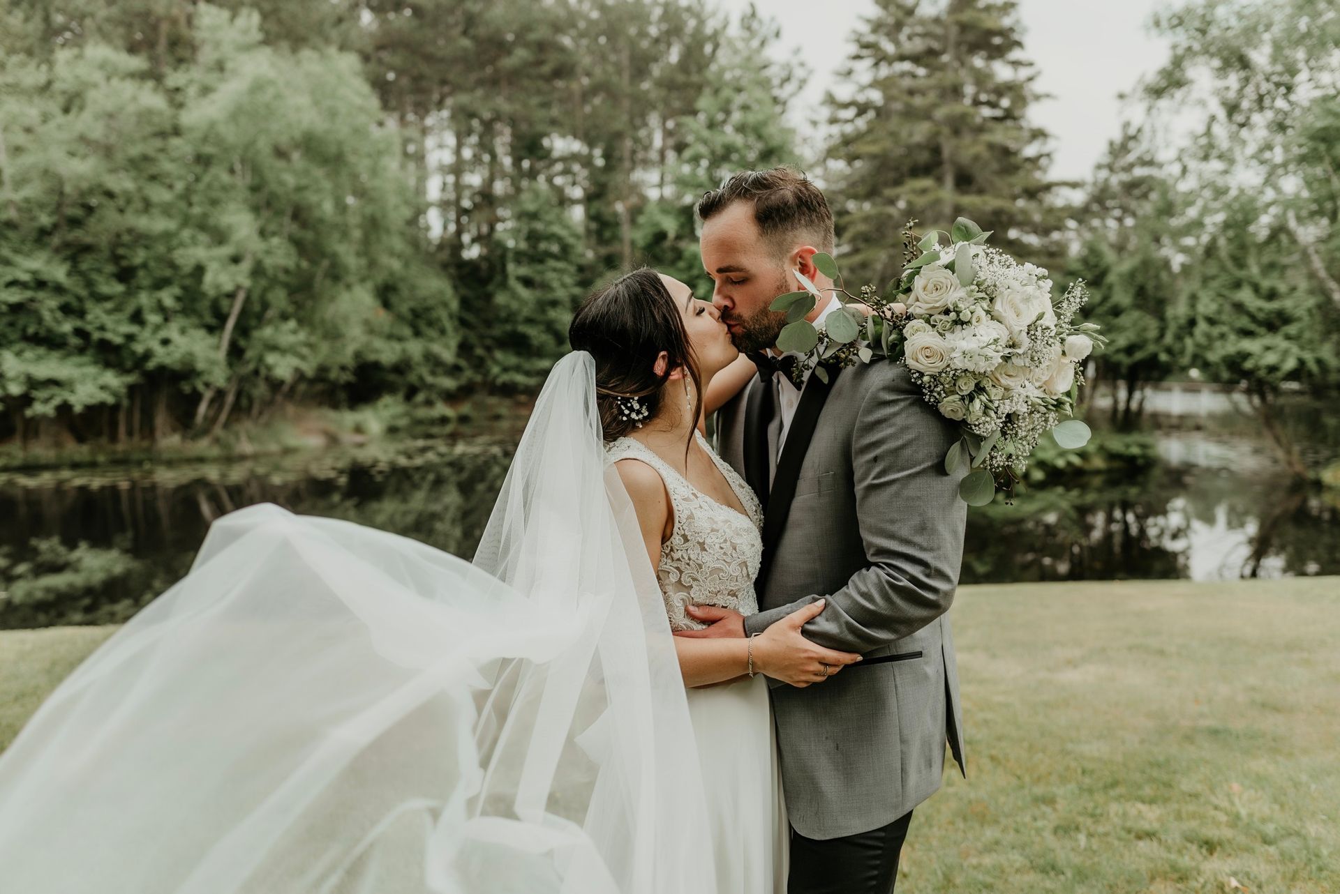 A bride and groom are kissing in front of a lake.