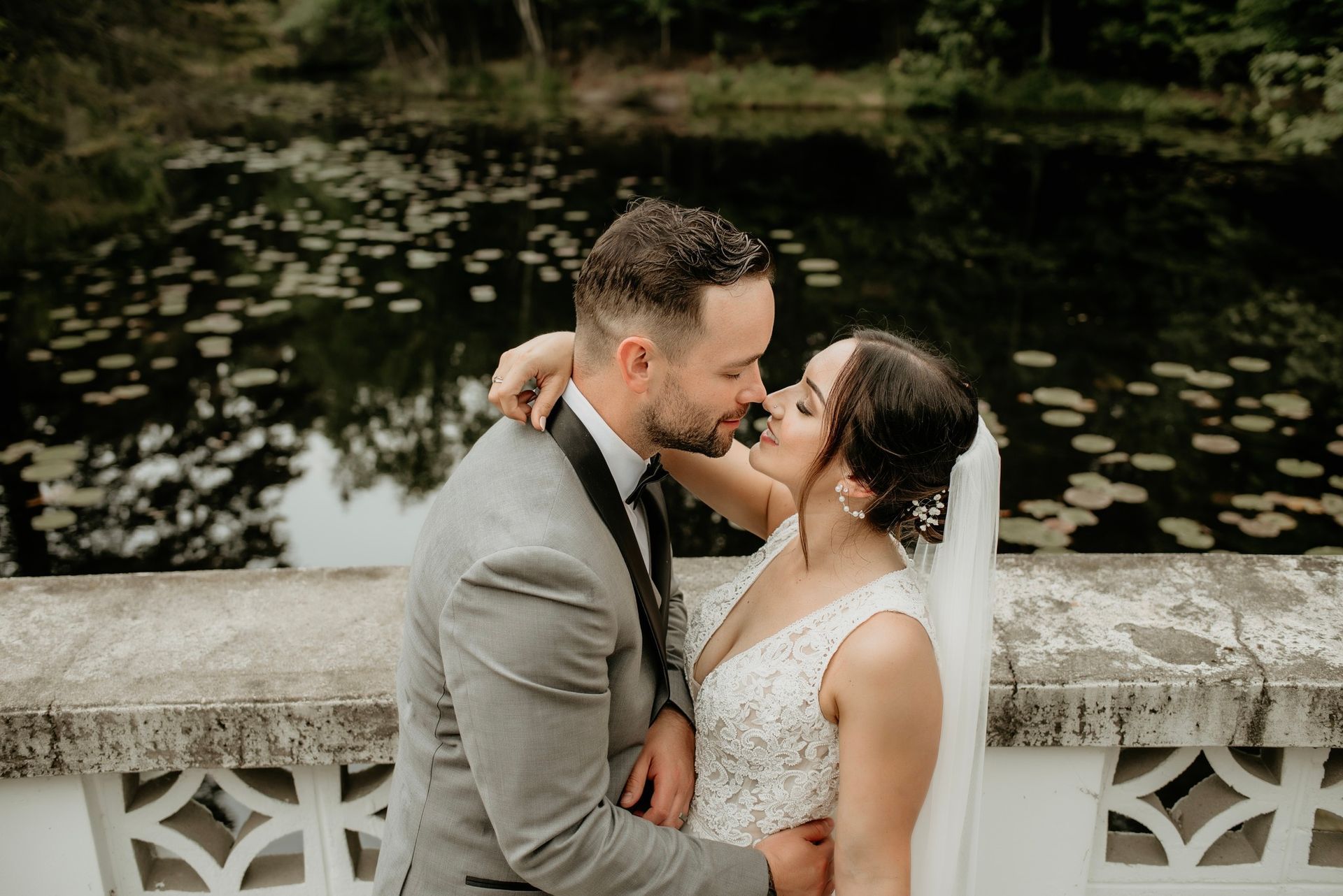 A bride and groom are kissing on a bridge in front of a pond.