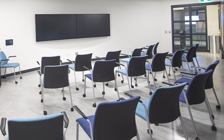 Rows of blue chairs face two large black screens in a modern classroom setting.