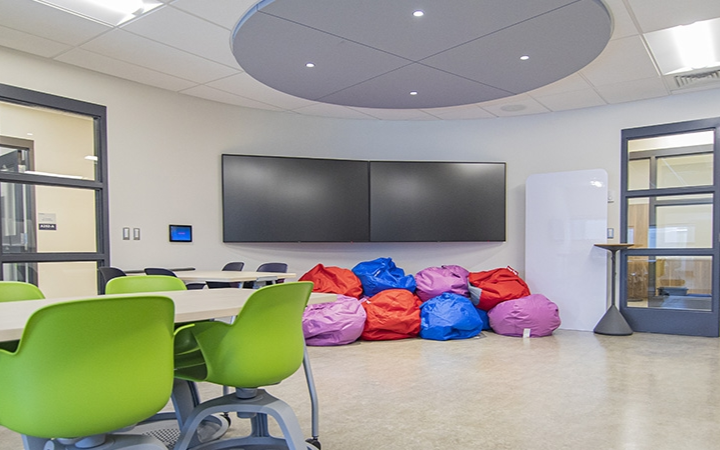 A modern classroom with green chairs, bean bags, large screens, and glass doors.