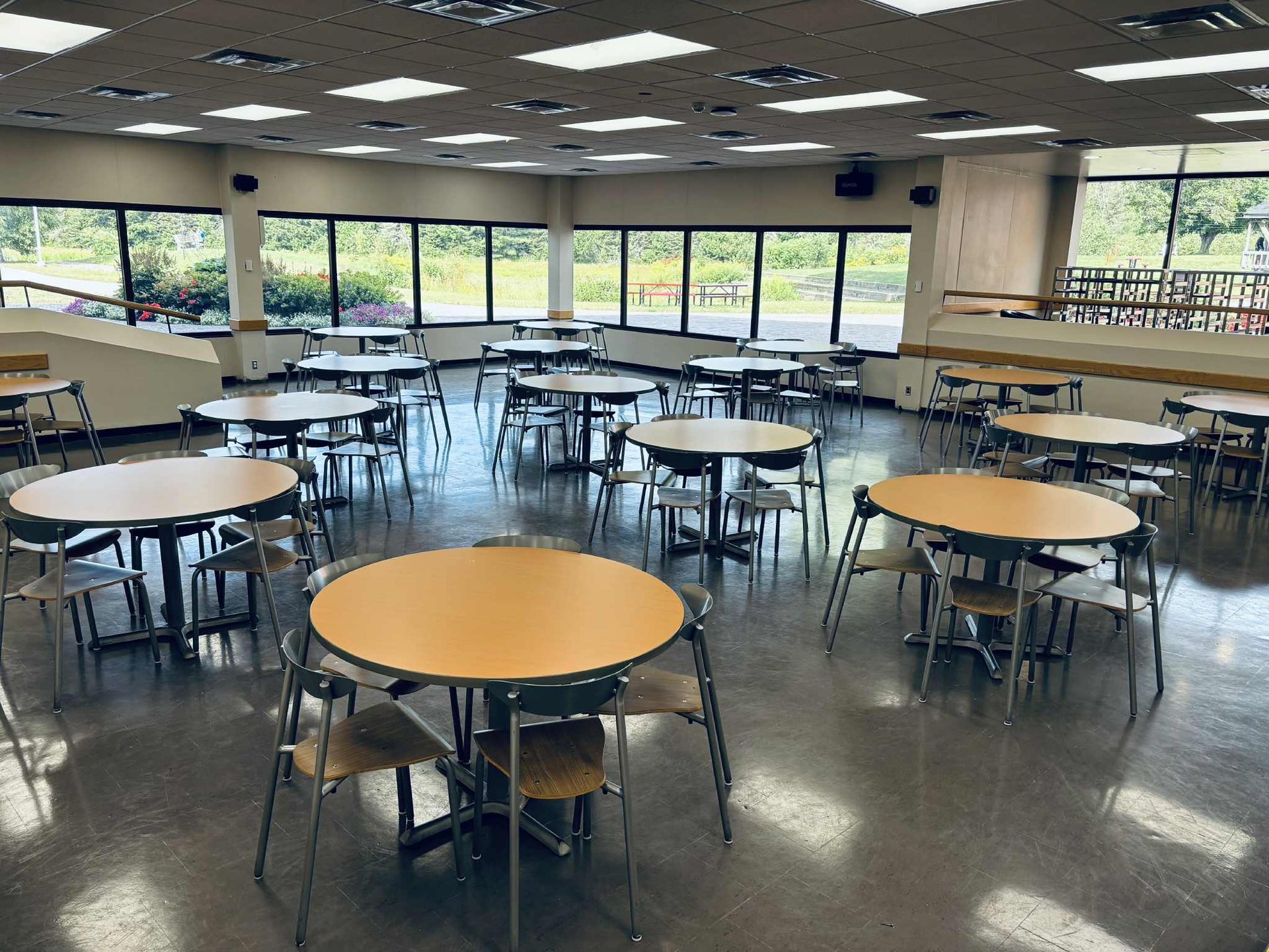 Empty cafeteria with round tables and chairs; large windows, bright lighting.