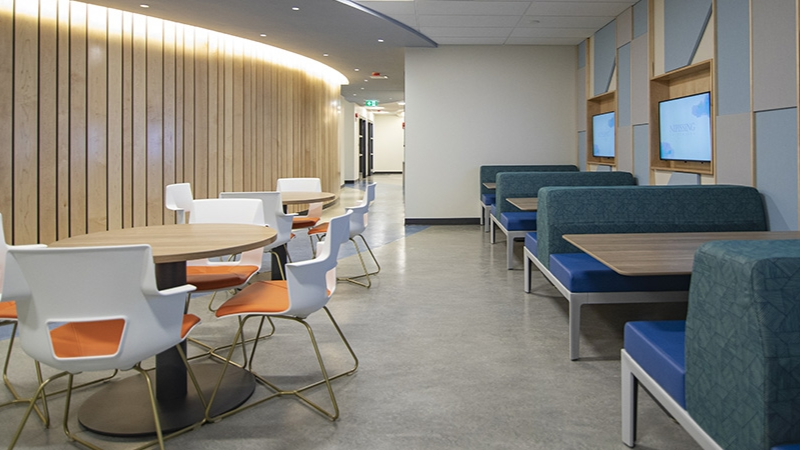 A modern cafeteria with tables, chairs, and booths; a curved wall with wood paneling and a neutral floor.