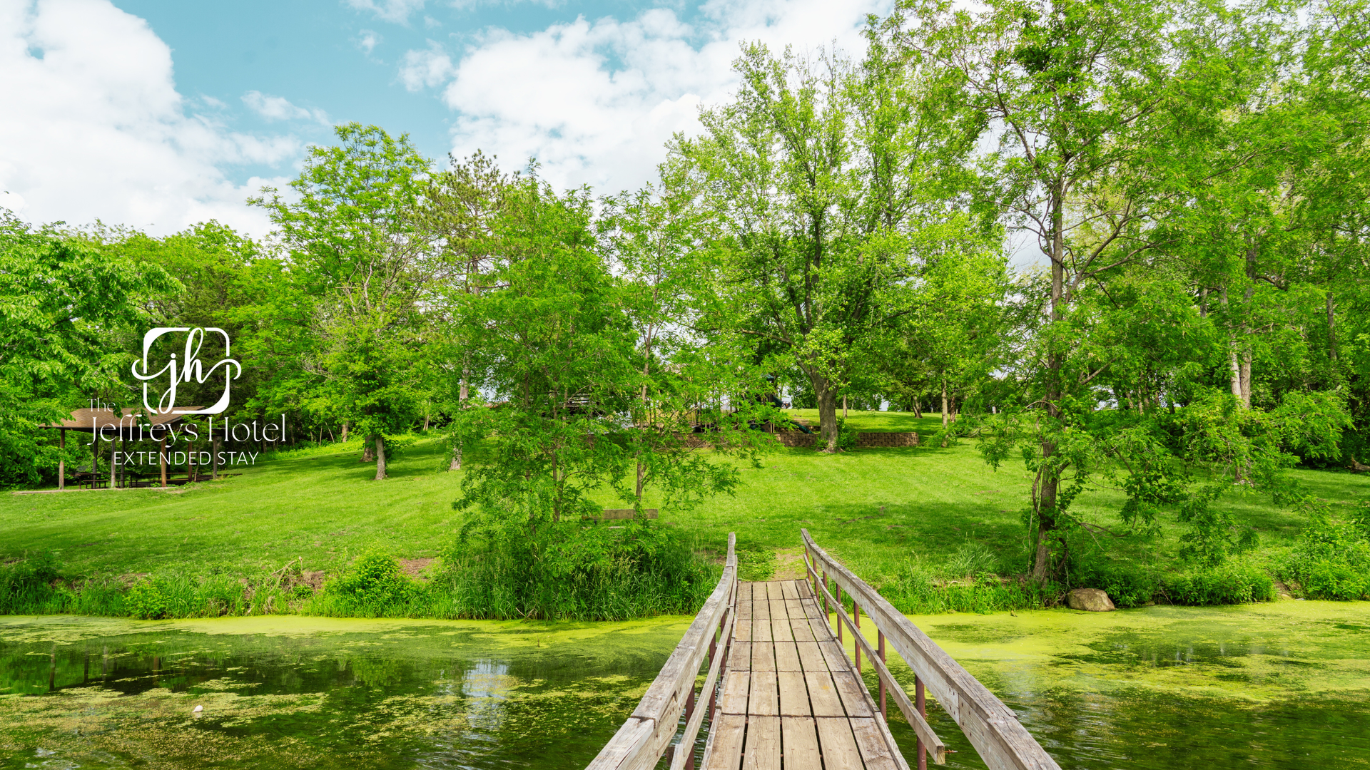 Wooden bridge over East Lake, Osceola, Iowa, leads to a grassy bank with trees under a cloudy sky. 