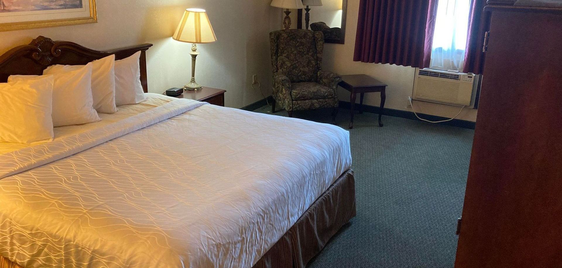 Hotel room with a king-sized bed, armchair, and lamp. Dark red curtains, and green carpet.