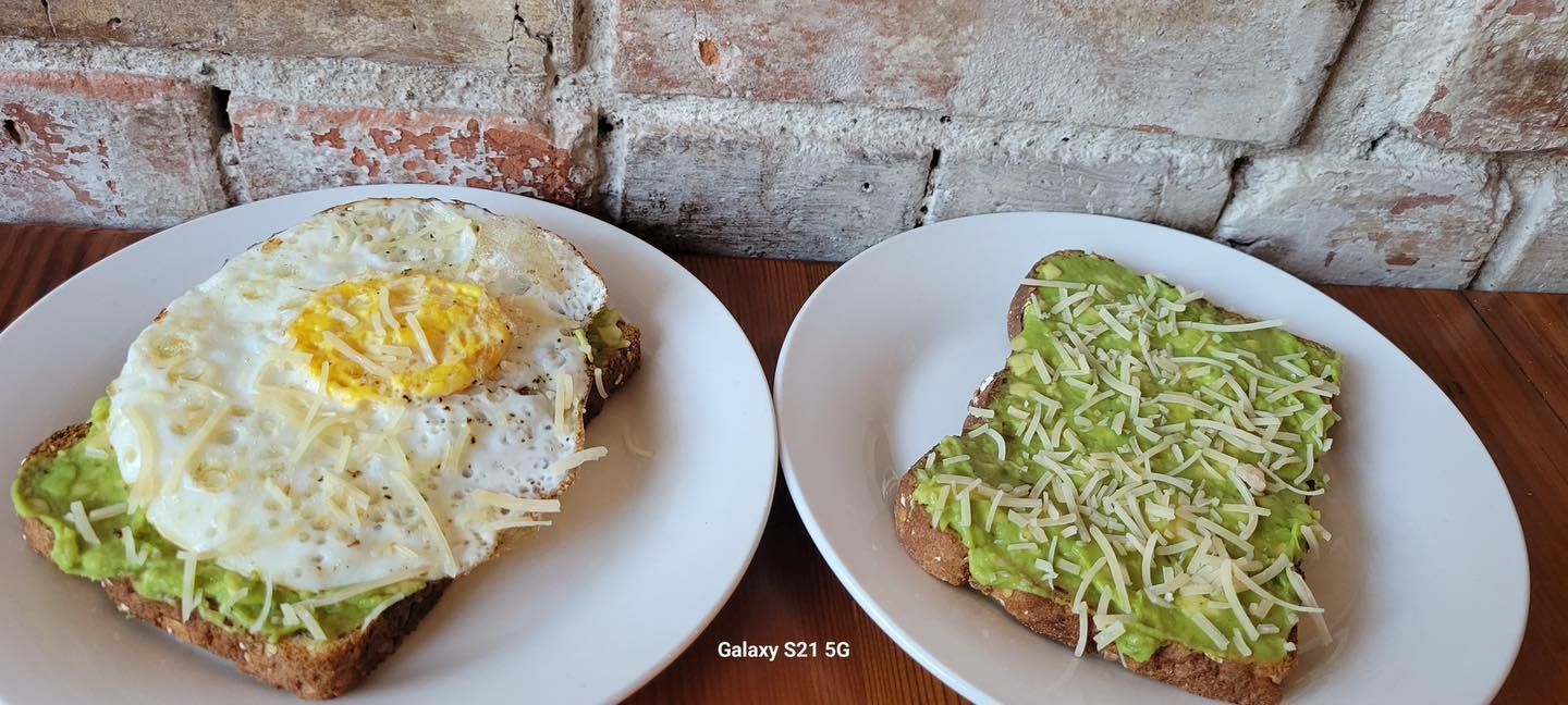 Two avocado toasts on white plates, one with an egg and one with shredded cheese, brick background.