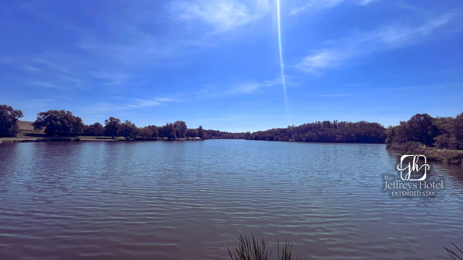 Blue lake under a bright blue sky, trees lining the far shore, bright sunburst.