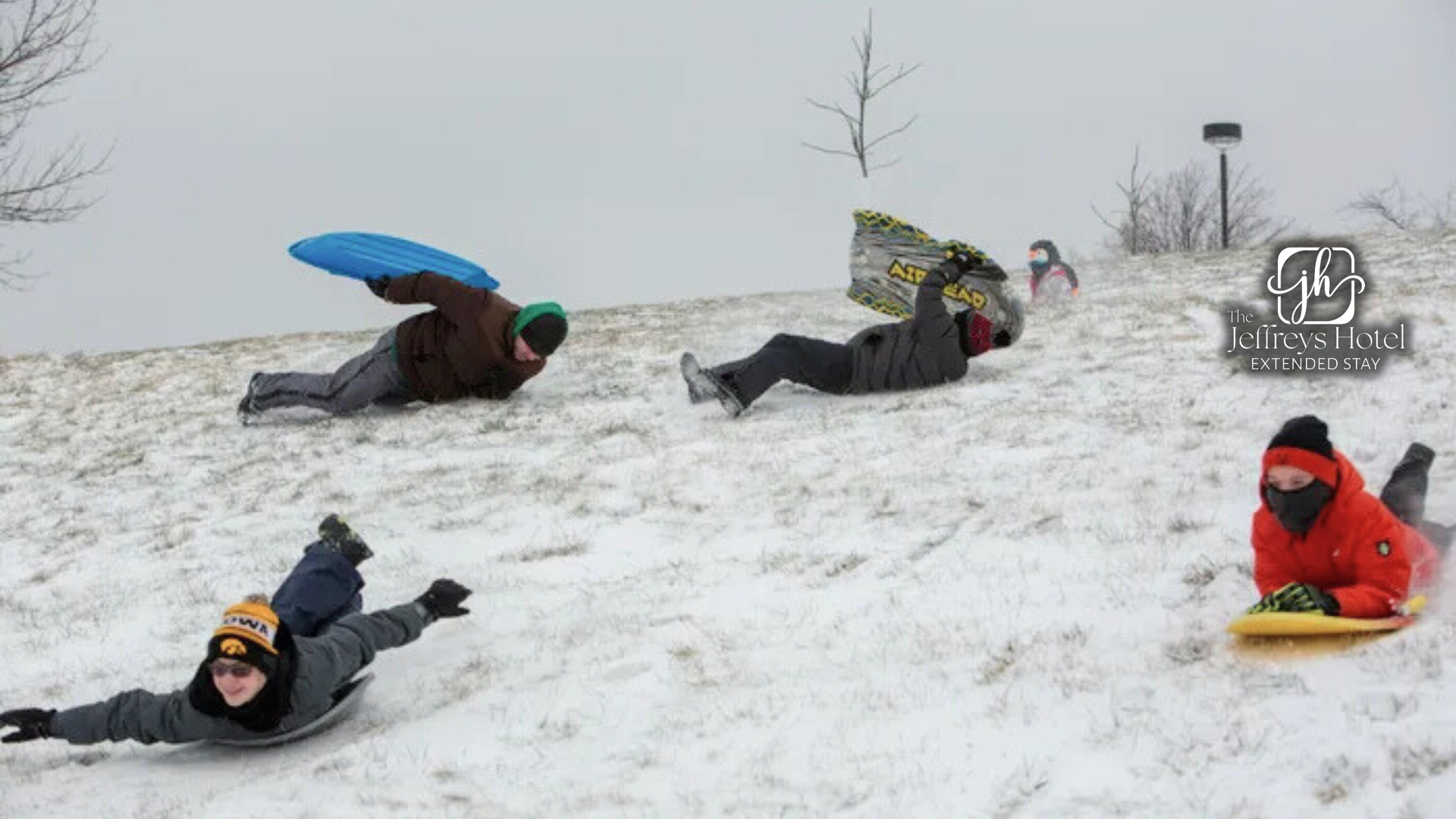 Children sledding down a snowy hill on saucers and sleds; overcast day.